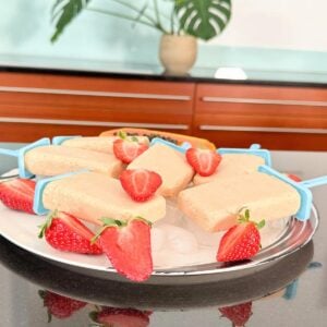 Creamy papaya strawberry popsicles on a plate with halved fresh strawberries, served on ice, with papaya and tropical plants in the background