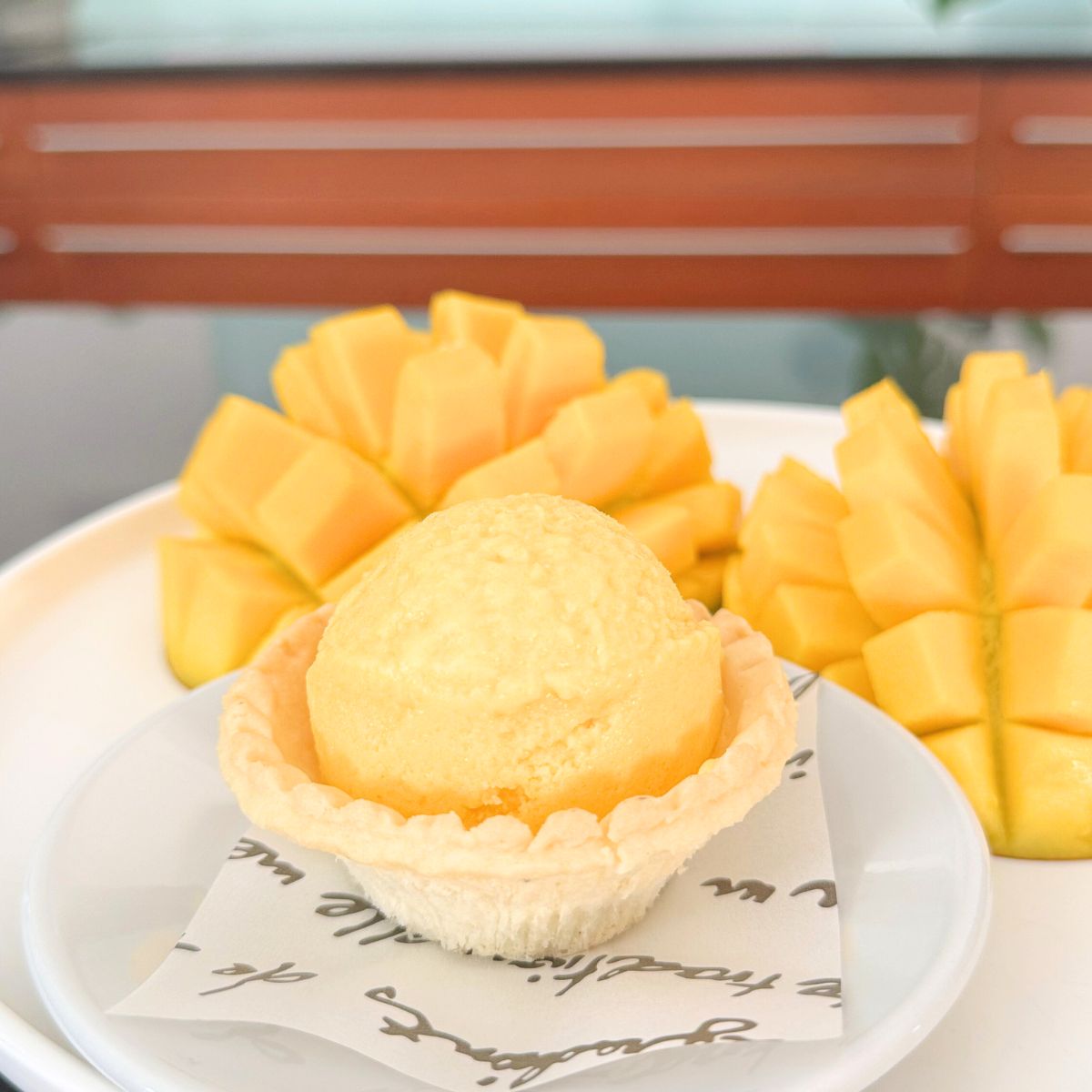 Mango frozen yogurt tart on a white plate with two halves of sliced fresh mango in the background, served on branded parchment paper.