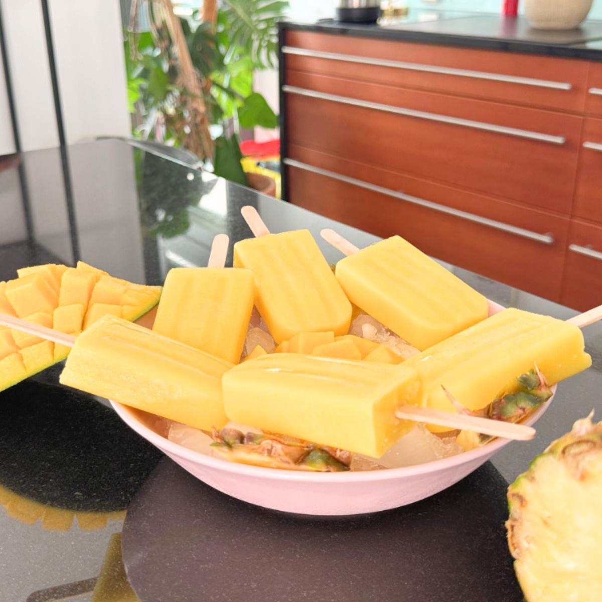 Bowl of pineapple-orange mango popsicles on ice, garnished with pineapple wedges and diced mango, set on a kitchen counter with plants in the background