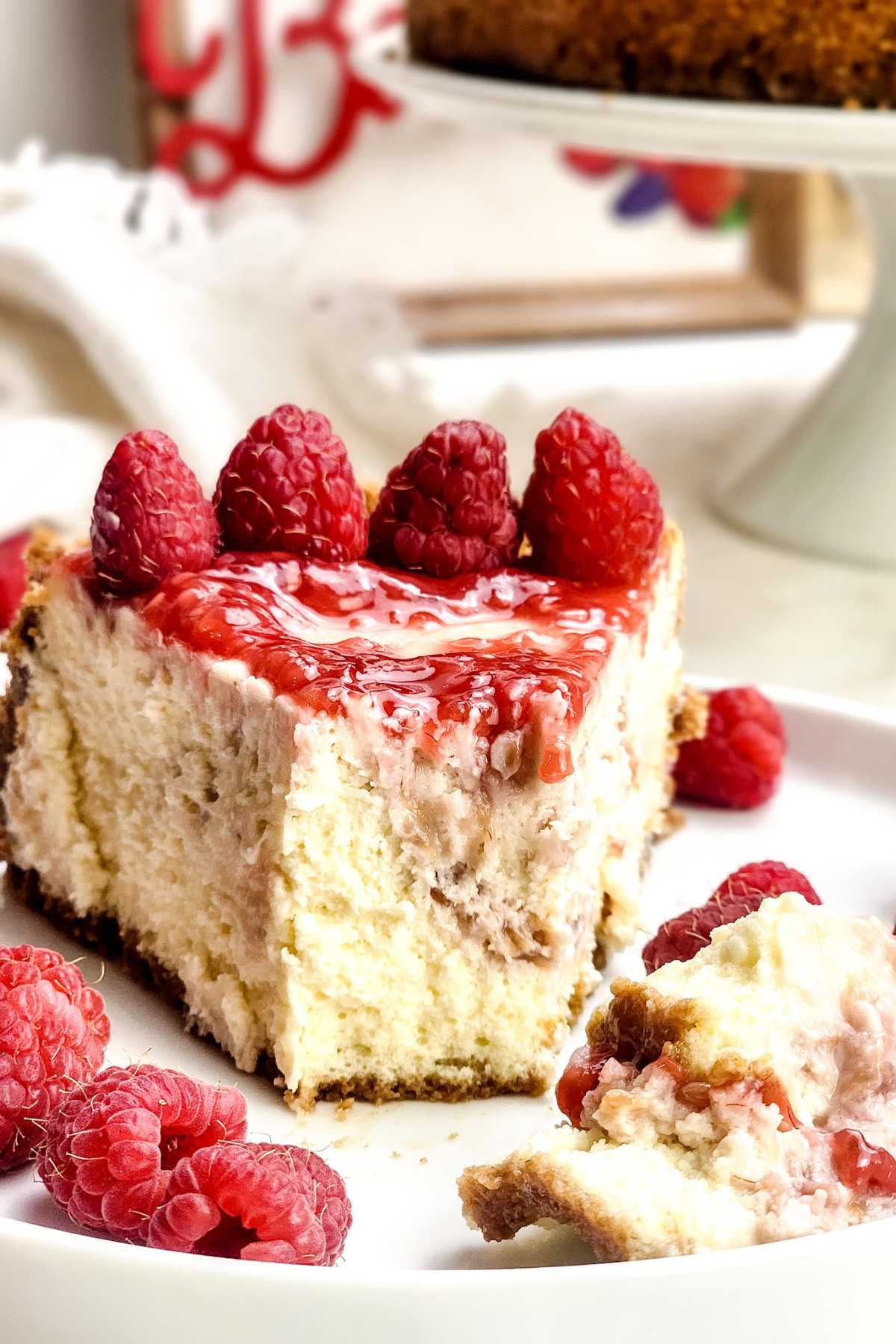 Close-up of a slice of raspberry cheesecake on a white plate, topped with glossy raspberry sauce and fresh raspberries. The creamy cheesecake filling and crumbly graham cracker crust are visible, with extra raspberries scattered around and a bite taken out on the plate.