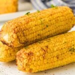 Close-up of three air fryer sweetcorn cobs stacked on a plate, lightly charred with seasoning and garnished with fresh herbs.