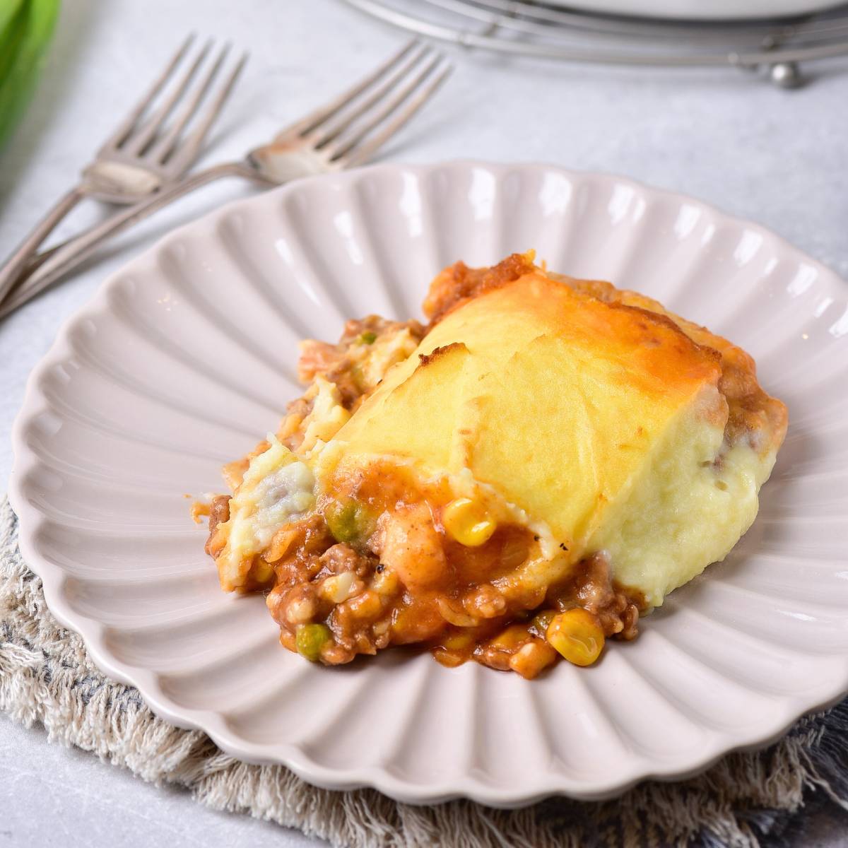 Close-up of a serving of shepherd’s pie on a scalloped beige plate, showing a golden-brown mashed potato topping with a hearty filling of ground meat, peas, corn, and carrots peeking out, photographed at an angle that highlights its layers and texture.