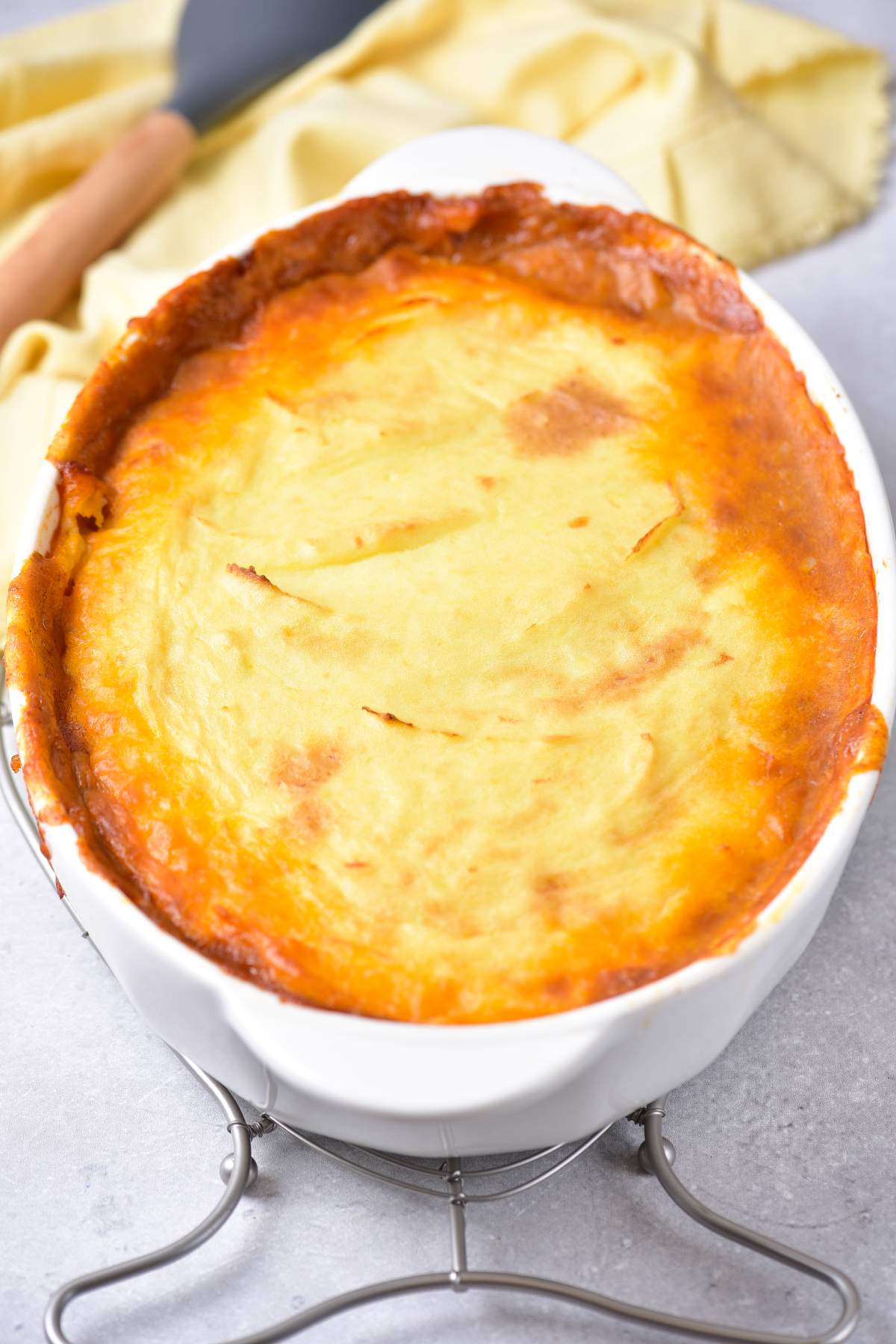 Baked shepherd&rsquo;s pie in a white oval dish, showing a golden-brown mashed potato topping with slightly crisp edges, resting on a metal trivet with a yellow cloth in the background.