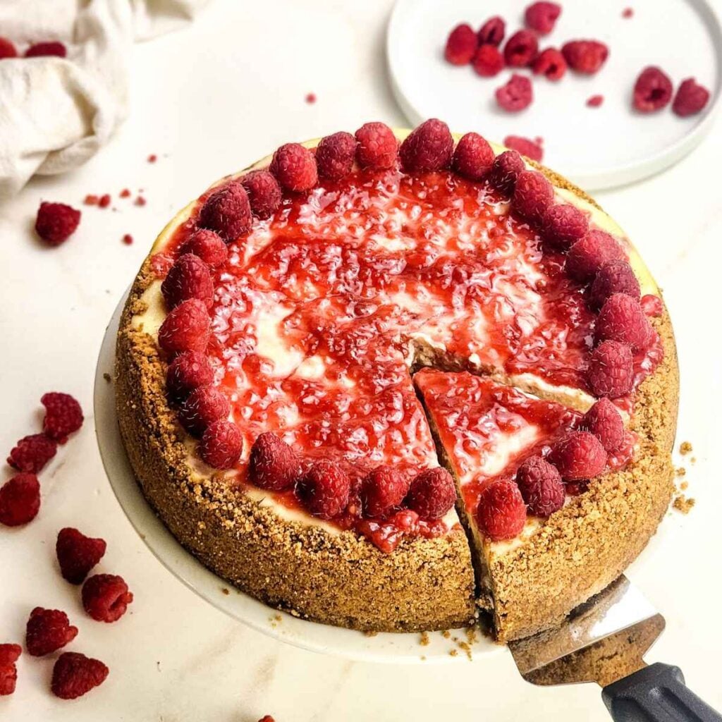 Tarta de queso con frambuesa entera con una base dorada de galleta Graham, cubierta con salsa de frambuesa brillante y frambuesas frescas alrededor del borde. Se est&aacute; levantando una porci&oacute;n, revelando el cremoso relleno de la tarta de queso, con frambuesas frescas esparcidas sobre la mesa al fondo.