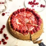 Whole raspberry cheesecake with a golden graham cracker crust, topped with glossy raspberry sauce and fresh raspberries around the edge. A slice is being lifted out, revealing the creamy cheesecake filling, with scattered fresh raspberries on the table in the background.