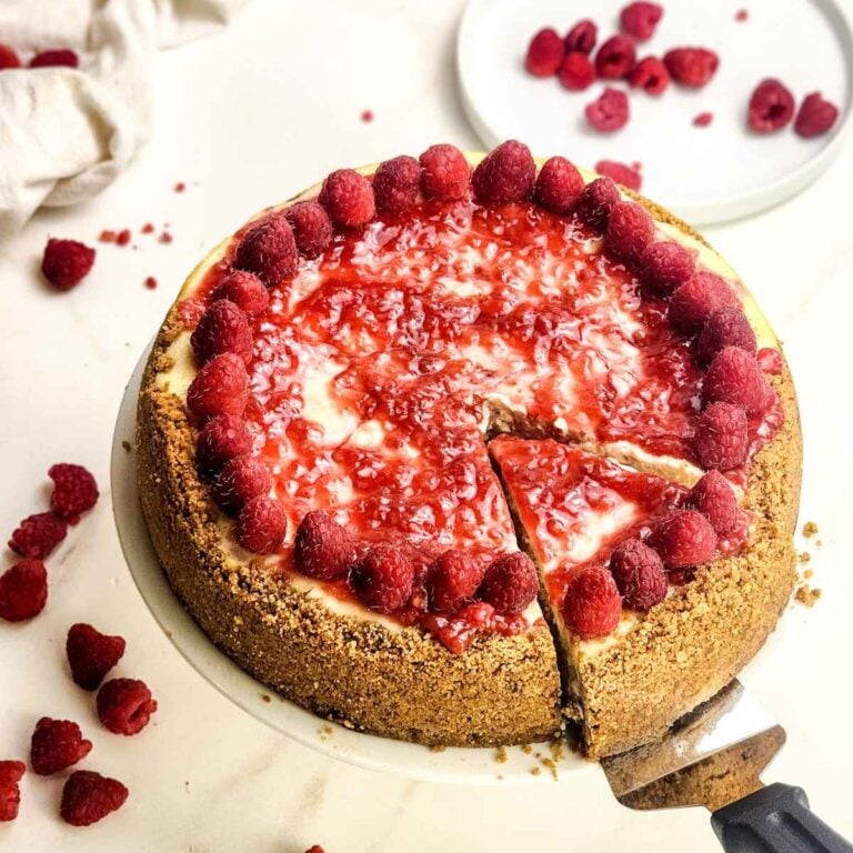 Whole raspberry cheesecake with a golden graham cracker crust, topped with glossy raspberry sauce and fresh raspberries around the edge. A slice is being lifted out, revealing the creamy cheesecake filling, with scattered fresh raspberries on the table in the background.