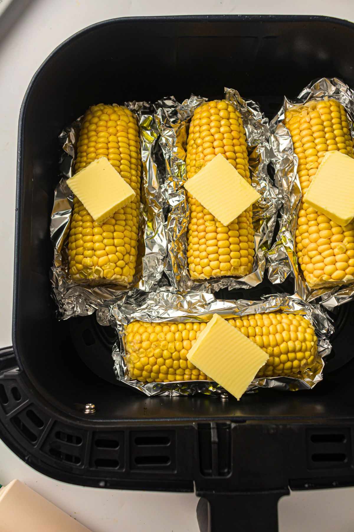 Four ears of corn wrapped in foil inside an air fryer basket, each topped with a square of butter before cooking.