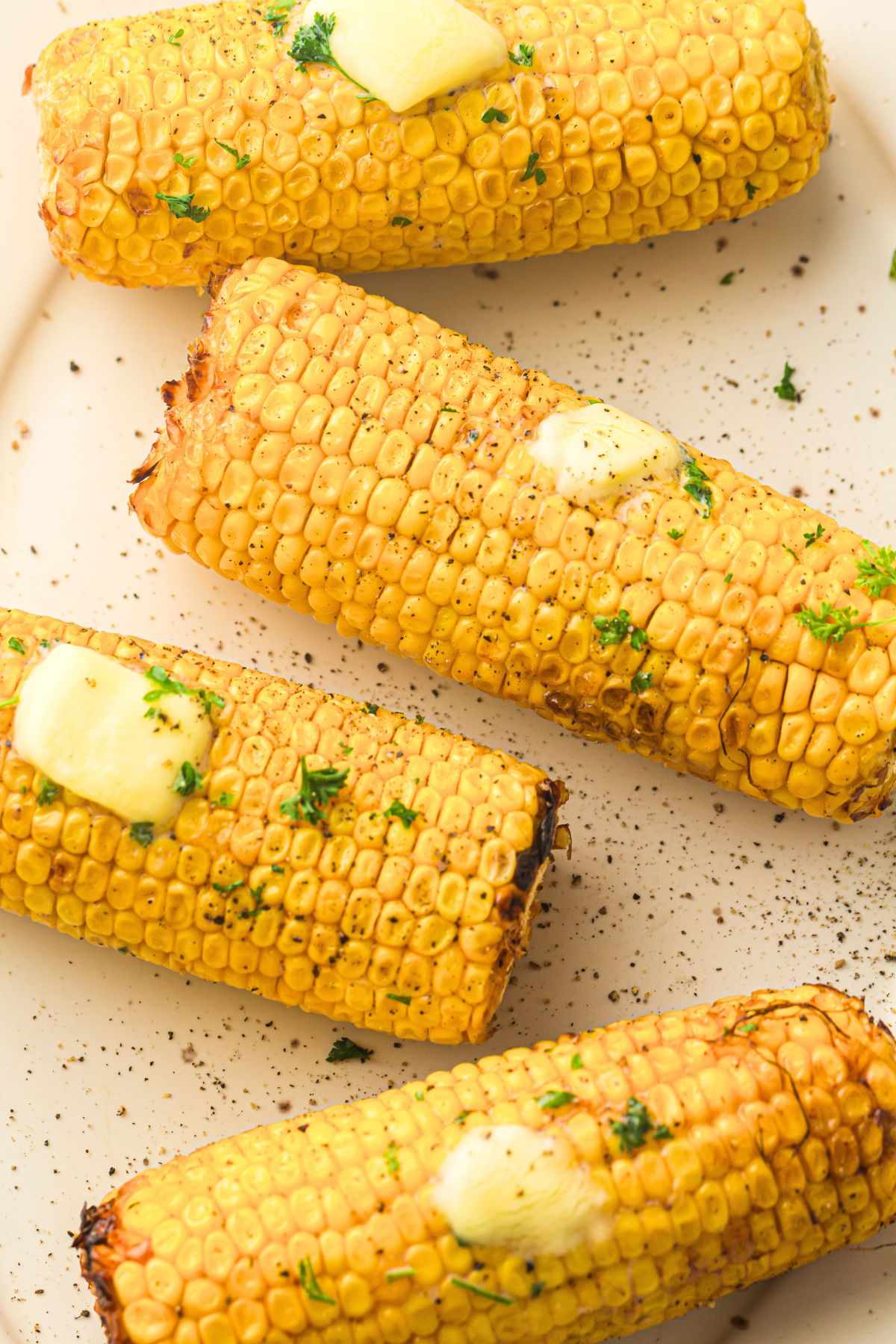 Four air fryer sweetcorn cobs on a plate, topped with melting butter and sprinkled with fresh herbs and black pepper.