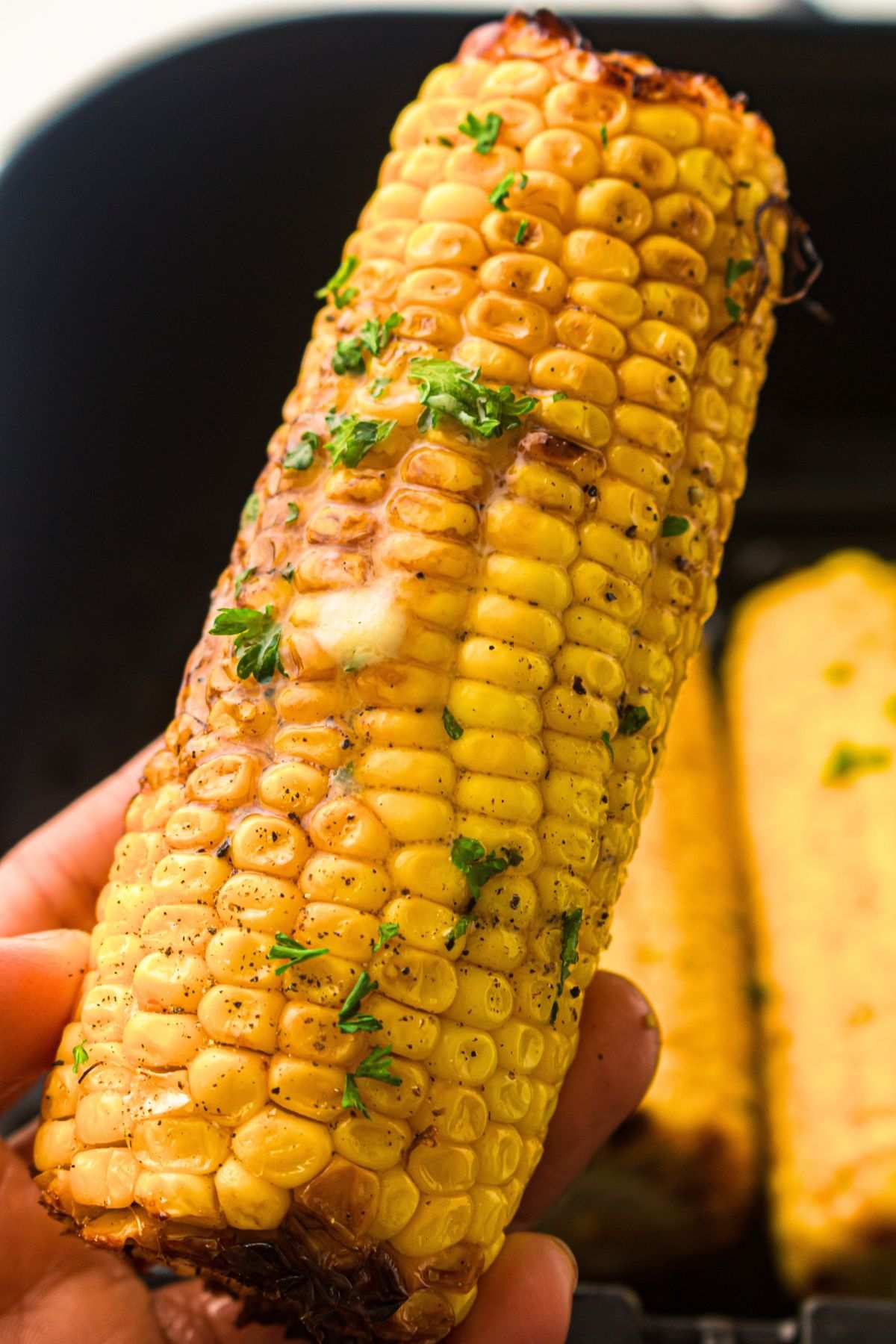 Hand holding a golden air fryer sweetcorn cob, glistening with melted butter, sprinkled with fresh herbs, and lightly charred on the edges.