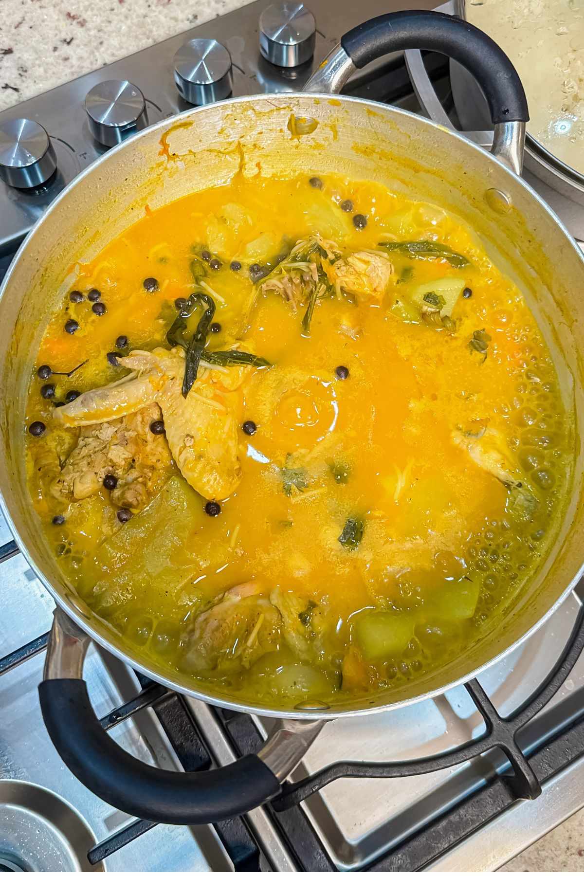 Dominican chicken soup cooking in a pot with chicken, golden broth, herbs, peppers, and whole spices.