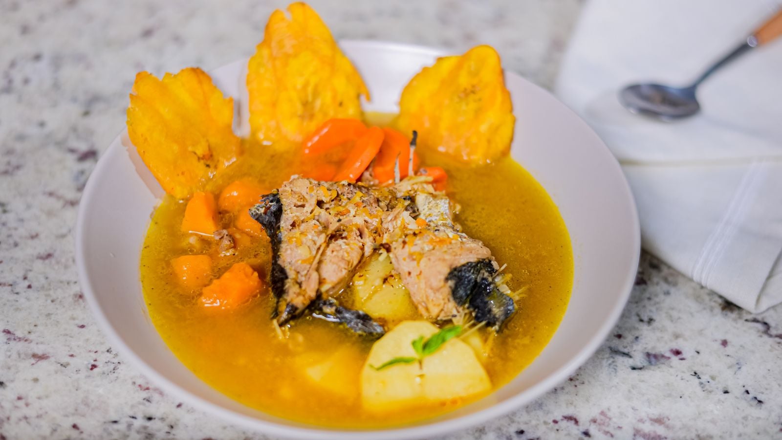 Dominican mahi-mahi fish soup served in a white bowl with vegetables, broth, and fried plantains.