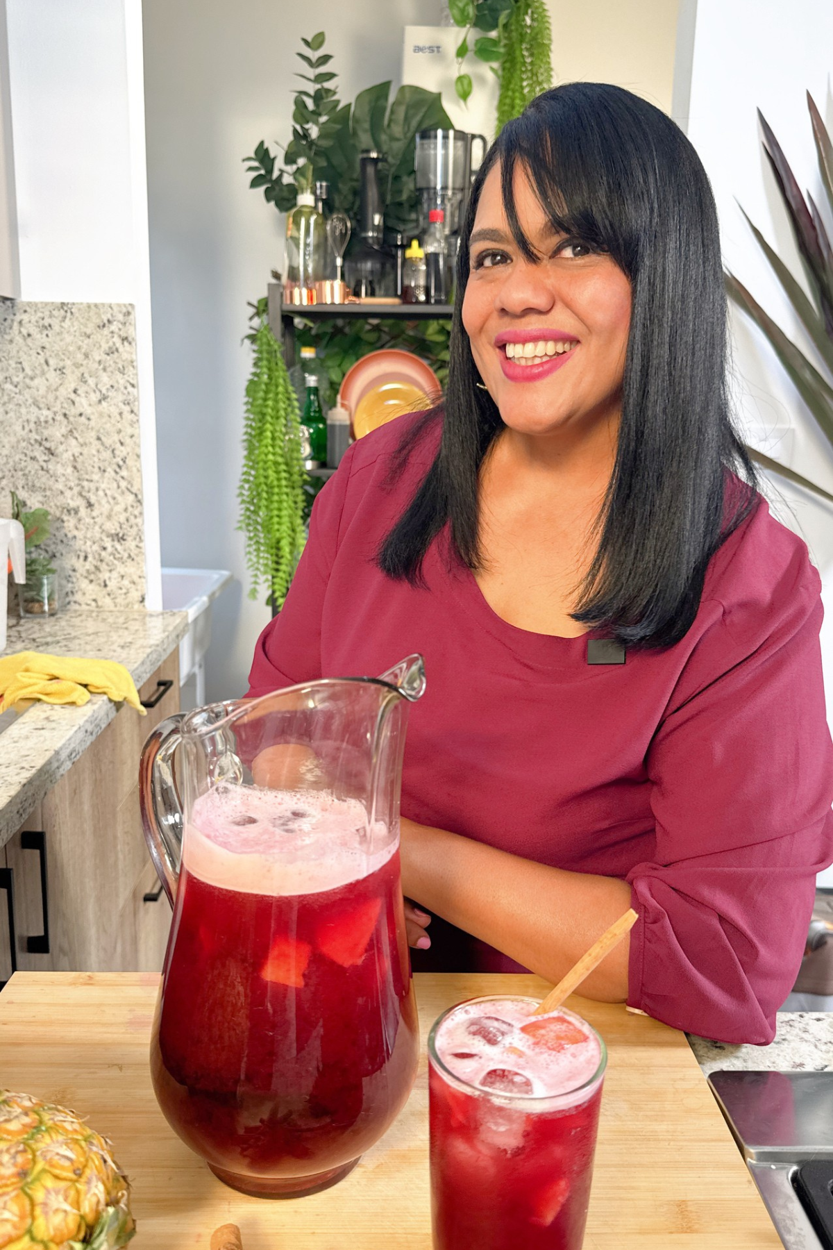 Mujer junto a una jarra y un vaso de jugo de piña con jamaica, con piña, canela y flores de jamaica sobre la mesa.