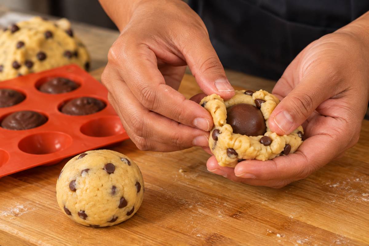 Hands folding chocolate chip cookie dough around a frozen hazelnut cream filling.